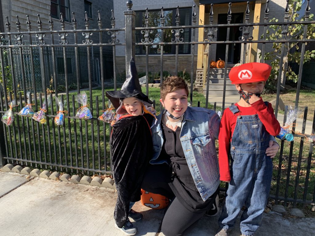 Michelle, smiling, flanked by her children. They are dressed up for Halloween as Mario and a witch.