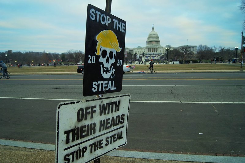 Two metal signs on a signpost. The upper is black and square, spray-painted with the words "Stop The Steal" framing a cigarette-smoking skull with ex-president Trump's signature orange toupee. The bottom sign is rectangular and spray-painted with the words "Off With Their Heads. Stop The Steal." (Blog Post: "Employment At Will" in Illinois)