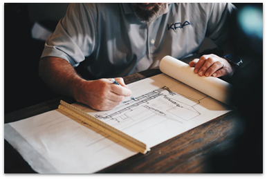 A man sits at a table, architectural drawings spread out in front of him. He is working on completing them.