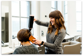 A smiling woman blowdries the hair of another woman, who sits in a salon chair with a black cape to keep hair trimmings off of her clothing.