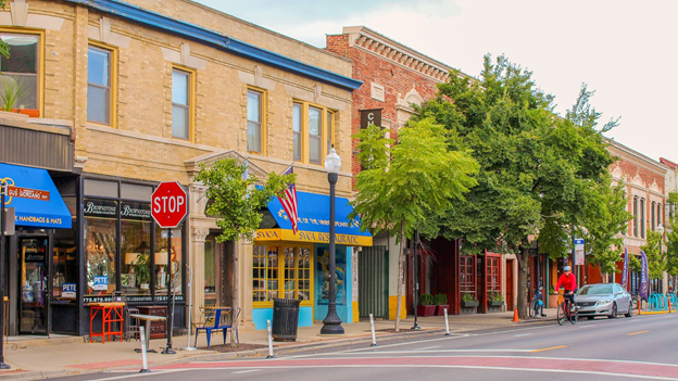 A row of storefronts on a tree-lined main street: one with a window full of antiques, one restaurant with a blue awning, and more businesses hidden behind trees.